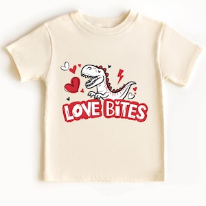 May include: Cream-colored t-shirt featuring a cartoon dinosaur with red accents, hearts, and a lightning bolt. The text "LOVE BITES" is printed in red below the dinosaur. A fun and playful design for kids.