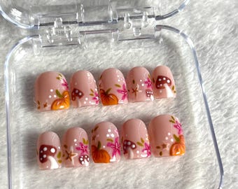 May include: A set of ten artificial nails in a clear plastic case. The nails are a light pink with autumn-themed designs, including orange pumpkins, brown mushrooms, and pink floral accents. The nails are oval-shaped.