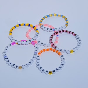 Op de afbeelding: Vijf kleurrijke kralenarmbanden met verschillende woorden erop. De armbanden zijn gemaakt van witte kralen en kralen in verschillende kleuren. De woorden op de armbanden zijn "GOOD GIRL", "SMUT", "WARNING", "COFFEE" en "BOOKS SPICY".
