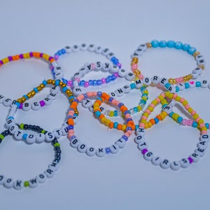 May include: A collection of colorful beaded bracelets with different words spelled out on them, including "Rather Be Reading", "Not Distracted", "Bookworm", "One More Book", "Hot Girls Read", and "Books".