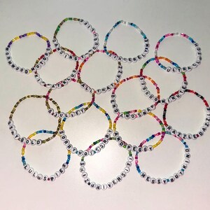 Puede incluir: Una colección de pulseras de cuentas con letras blancas que deletrean varias palabras y frases. Las pulseras presentan cuentas coloridas en tonos arcoíris. Las palabras incluyen "Sorry", "Messy", "Kind" y "Dazzle".