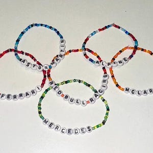 May include: Six colorful beaded bracelets with the names of Formula 1 racing teams: Ferrari, Aston Martin, Alpine, McLaren, Red Bull Racing, and Mercedes.