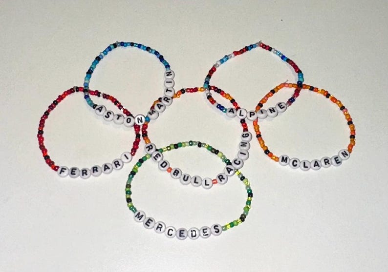 May include: Six colorful beaded bracelets with the names of Formula 1 racing teams: Ferrari, Aston Martin, Alpine, McLaren, Red Bull Racing, and Mercedes.