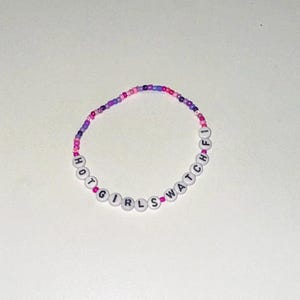 May include: A beaded bracelet with the words "HOT GIRLS WATCH" spelled out in black letters on white beads. The beads are interspersed with pink and purple beads.