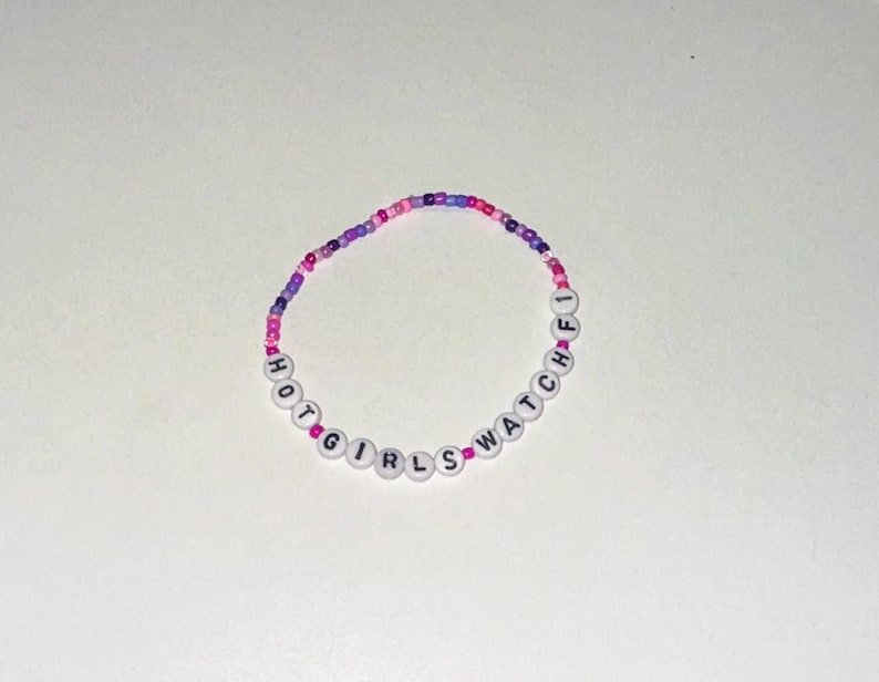 May include: A beaded bracelet with the words "HOT GIRLS WATCH" spelled out in black letters on white beads. The beads are interspersed with pink and purple beads.