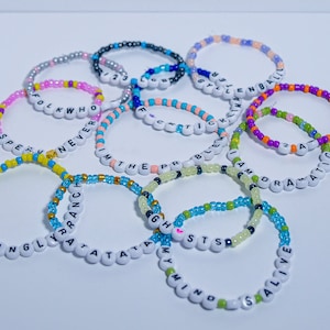 May include: A collection of colorful beaded bracelets with different words spelled out on them, including "Seemingly", "Y'all", "Tatas", "G.H.S.T.S", "Mind", "Stay Alive", "Folk Who", "Never", "With The", "Problem", "I Am", "Dramatic", "Style", "Kim", "Blen", "Bali", "2 A".