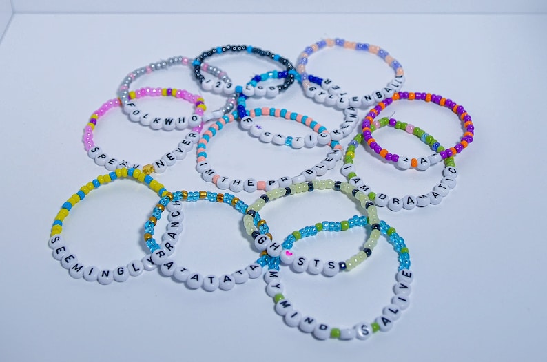 May include: A collection of colorful beaded bracelets with different words spelled out on them, including "Seemingly", "Y'all", "Tatas", "G.H.S.T.S", "Mind", "Stay Alive", "Folk Who", "Never", "With The", "Problem", "I Am", "Dramatic", "Style", "Kim", "Blen", "Bali", "2 A".