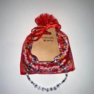 May include: A red mesh bag filled with colorful beaded bracelets. The bag has a brown tag that says "Handmade with love". A white beaded bracelet with the words "PIT CREW" is displayed in front of the bag.