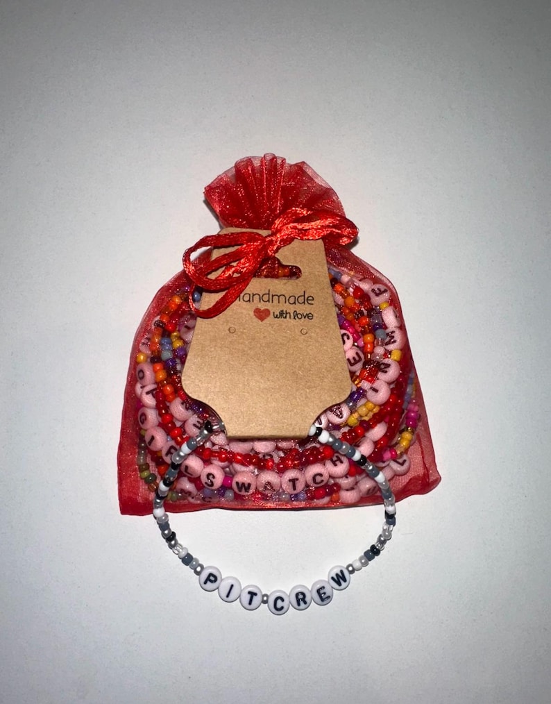 May include: A red mesh bag filled with colorful beaded bracelets. The bag has a brown tag that says "Handmade with love". A white beaded bracelet with the words "PIT CREW" is displayed in front of the bag.