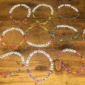May include: A collection of beaded bracelets with white letter beads spelling out phrases like "Magical," "Disneyland," and "Happily Ever After." The bracelets feature colorful beads in various hues, including pink, orange, and blue.