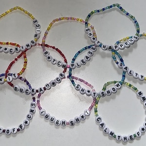 May include: A collection of beaded bracelets with white letter beads spelling out various phrases. The bracelets are in a variety of colors, including yellow, red, purple, blue, and green. The phrases include "BE F*CKING NICE", "F*CK IT", and "CALM THE F*CK DOWN".