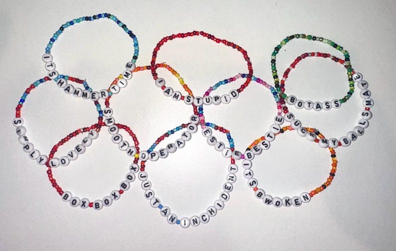 May include: A set of eight colorful beaded bracelets with different phrases spelled out on white beads. The phrases include "It's Hammer Time", "Simply Lovely", "Smooth Operator", "I Am Stupid", "Best Friends", "I'm The Best", "B.O.X.B.O.X", and "Botass My Balls Mate".