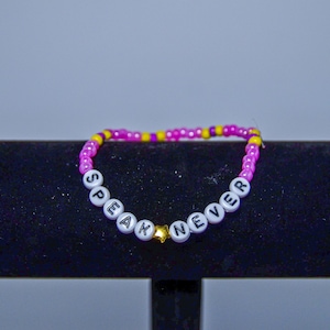 May include: Pink and yellow beaded bracelet with white letter beads spelling "Speak Never" with a gold star charm.
