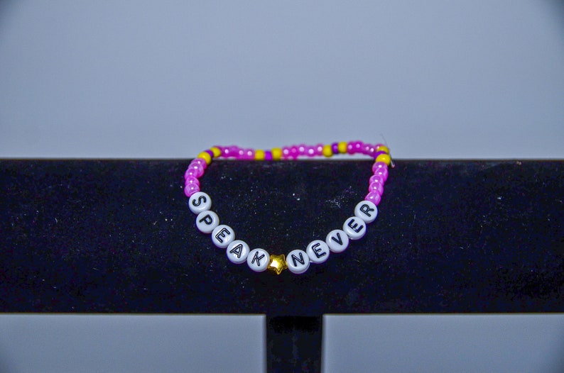 May include: Pink and yellow beaded bracelet with white letter beads spelling "Speak Never" with a gold star charm.