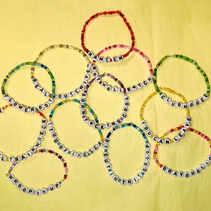 May include: A collection of colorful beaded bracelets with different phrases spelled out on them, including "Flowerbomb", "Not Well Bitch", "You Can Leave", and "Clubhouse".