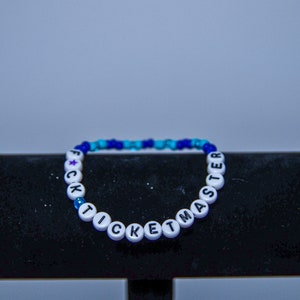 May include: A white beaded bracelet with the phrase "F*CK TICKETMASTER" spelled out in black letters. The beads are interspersed with blue and turquoise beads.
