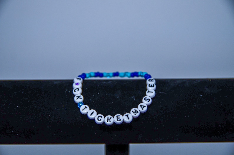 May include: A white beaded bracelet with the phrase "F*CK TICKETMASTER" spelled out in black letters. The beads are interspersed with blue and turquoise beads.