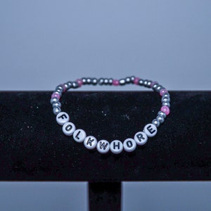 May include: A silver and pink beaded bracelet with the word "FOLKWHORE" spelled out in black letters on white beads.