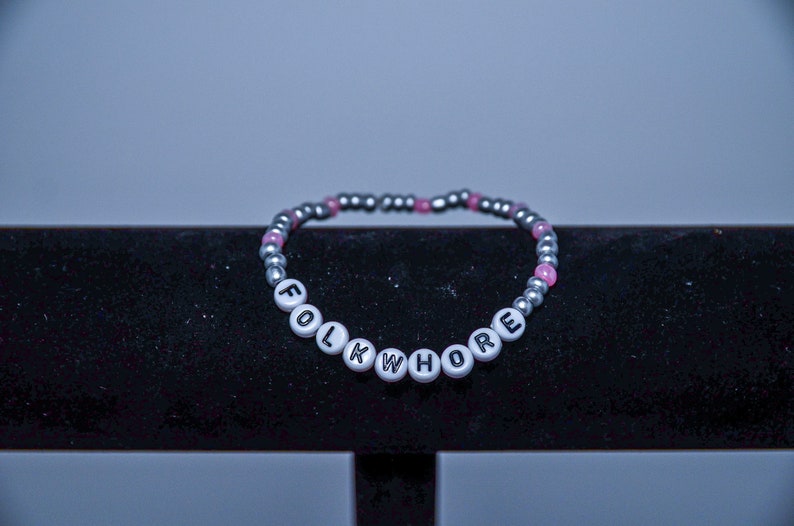 May include: A silver and pink beaded bracelet with the word "FOLKWHORE" spelled out in black letters on white beads.