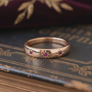 Puede incluir: Un anillo de oro con tres diseños de estrellas, cada uno con una pequeña gema roja redonda. El anillo se presenta sobre un fondo oscuro, resaltando su acabado pulido y sus intrincados detalles. Una pieza de joyería clásica.