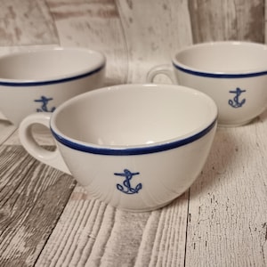 Könnte beinhalten: Drei weiße Teetassen aus Keramik mit blauem Rand und Anker-Design. Die Tassen stehen auf einer hellen Holzoberfläche.