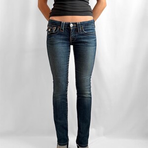 May include: Dark wash denim jeans with a classic five-pocket design. The jeans have a button and zipper closure, and are worn with a black top and dark shoes. The jeans are form-fitting.