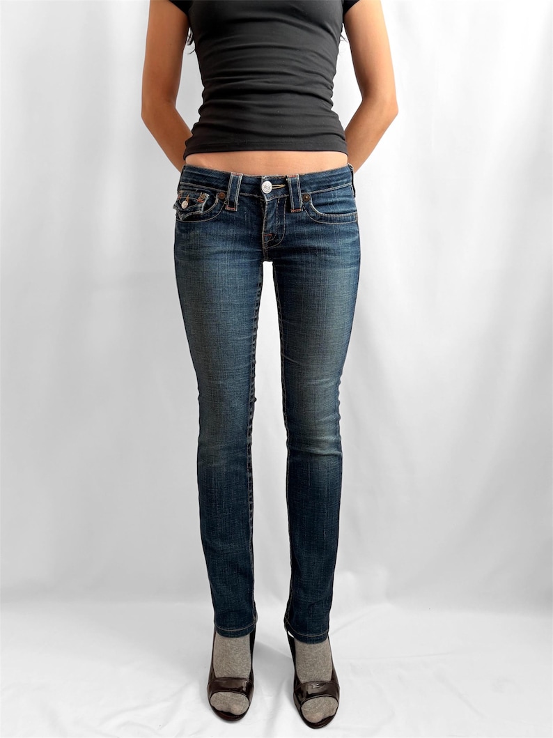 May include: Dark wash denim jeans with a classic five-pocket design. The jeans have a button and zipper closure, and are worn with a black top and dark shoes. The jeans are form-fitting.