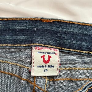 May include: Close-up of a pair of blue denim jeans. The label reads "BRAND JEANS" with a red horseshoe logo and "made in USA 24". The jeans have orange stitching and are on a white background.
