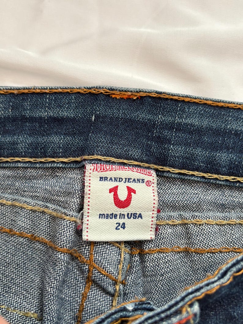May include: Close-up of a pair of blue denim jeans. The label reads "BRAND JEANS" with a red horseshoe logo and "made in USA 24". The jeans have orange stitching and are on a white background.