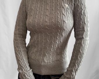 May include: A long-sleeved grey cable knit jumper featuring a crew neckline and a small white embroidered logo on the upper left chest. The garment has ribbed cuffs and a ribbed hem, providing a fitted silhouette with a classic textured pattern.