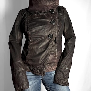 May include: A dark brown jacket with a high funnel collar and an asymmetrical front closure featuring three large black buttons. The jacket has long sleeves, a textured finish, and a fitted silhouette, worn over blue denim jeans.