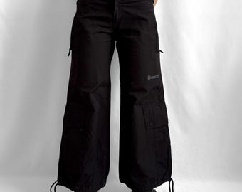 May include: Black cargo trousers featuring a wide-leg silhouette, multiple utility pockets on the thighs, and adjustable drawstring hems. The word Bench is printed in a subtle grey font on the left thigh area, and the waistband includes side adjustment tabs.