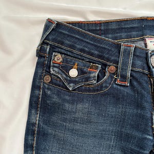 May include: Close-up of dark blue denim jeans. The jeans feature a classic five-pocket design with metal rivets and a button closure. Orange stitching accents the seams and pockets. The fabric has a slightly faded appearance.