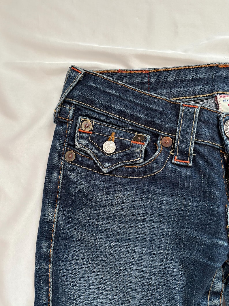 May include: Close-up of dark blue denim jeans. The jeans feature a classic five-pocket design with metal rivets and a button closure. Orange stitching accents the seams and pockets. The fabric has a slightly faded appearance.