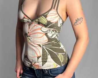 May include: A sleeveless vest top featuring a deep V-neckline and double spaghetti straps. The fabric is patterned with large, stylised peach and white flowers against an olive green background, paired with dark blue denim jeans.