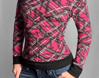 May include: A long-sleeve off-the-shoulder top featuring a vibrant pink and black plaid pattern with white intersecting lines and black script text overlays. The garment includes a solid black wide neckline trim, black cuffs, and a black hem.