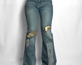 May include: Blue denim jeans featuring a mid-rise waist, a button and zip fly, and a flared leg silhouette. The jeans are decorated with gold metallic fabric patches, embroidered floral details, and a large vertical panel on the lower leg.
