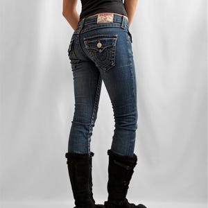May include: Blue denim jeans with a faded wash, featuring a decorative back pocket design and a leather patch. The jeans are paired with black knee-high boots with faux fur trim. A black tank top completes the outfit.