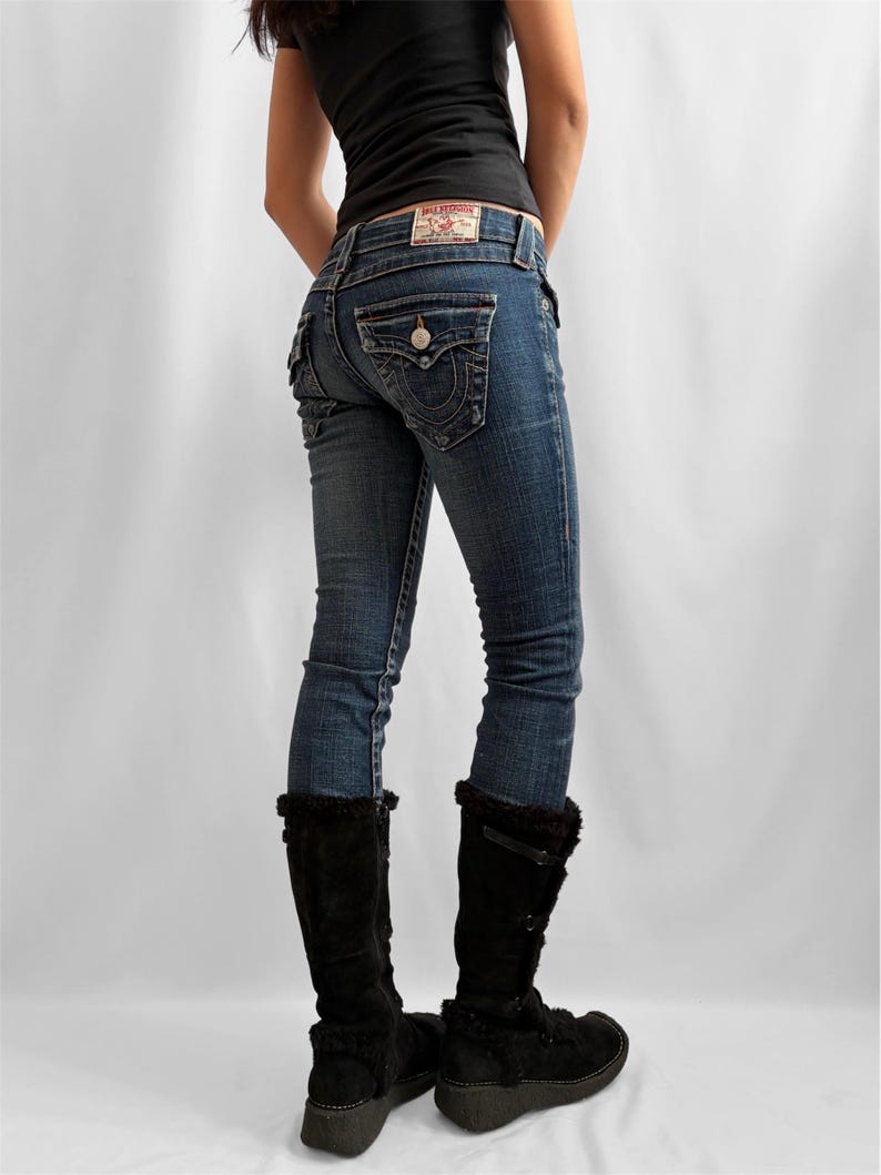 May include: Blue denim jeans with a faded wash, featuring a decorative back pocket design and a leather patch. The jeans are paired with black knee-high boots with faux fur trim. A black tank top completes the outfit.