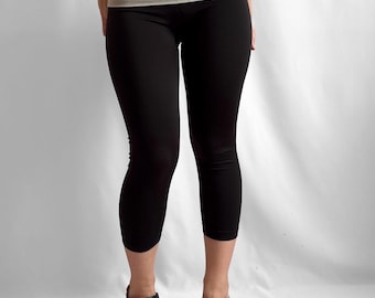 May include: Black cropped leggings featuring a wide, fold-over light grey waistband with the word PINK printed in black lettering. The leggings are paired with black leather clogs that have wooden soles and a rounded toe design.