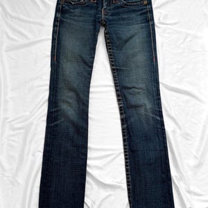May include: Dark wash denim jeans with a classic five-pocket design. The jeans feature a button and zipper closure, with orange stitching details along the seams and pockets. The jeans are laid flat on a white surface.