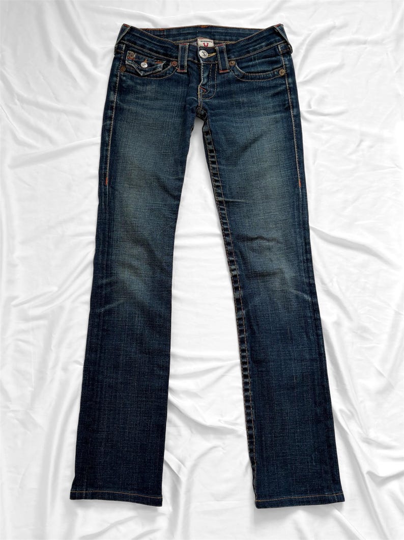 May include: Dark wash denim jeans with a classic five-pocket design. The jeans feature a button and zipper closure, with orange stitching details along the seams and pockets. The jeans are laid flat on a white surface.
