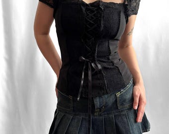 May include: A black short-sleeved top featuring lace panels, a square neckline, and a central corset-style lace-up closure with a ribbon tie, paired with a dark blue denim pleated mini skirt and bright pink and green leg warmers.