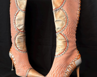 May include: A pair of knee-high boots featuring a peach-coloured snakeskin-textured pattern, metallic silver panels, and orange lace-up detailing along the sides. These pointed-toe boots include a stiletto heel and decorative silver studs.