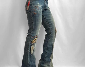 May include: Blue denim jeans featuring a flared leg silhouette, distressed detailing, and decorative floral embroidery. The jeans include a red brand tag on the hip, patchwork accents on the lower legs, and a medium wash finish with fading.