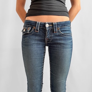 May include: Dark wash denim jeans with a button and zipper closure, featuring classic five-pocket styling. The jeans have a slim fit and are paired with a black tank top. The jeans have red stitching details and a small pocket on the front.