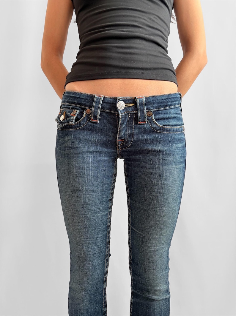 May include: Dark wash denim jeans with a button and zipper closure, featuring classic five-pocket styling. The jeans have a slim fit and are paired with a black tank top. The jeans have red stitching details and a small pocket on the front.