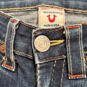 May include: Close-up of dark blue denim jeans. The jeans feature a button closure with the text "BRAND JEANS TRUE RELIGION" and a red and white label that says "BRAND JEANS" and "made in USA". Orange stitching accents the seams.