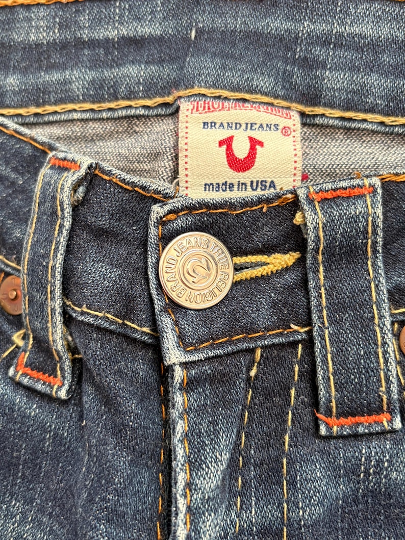 May include: Close-up of dark blue denim jeans. The jeans feature a button closure with the text "BRAND JEANS TRUE RELIGION" and a red and white label that says "BRAND JEANS" and "made in USA". Orange stitching accents the seams.