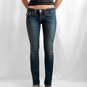 May include: Dark wash denim jeans with a classic five-pocket design. The jeans have a button and zipper closure, and are worn with a black tank top and dark shoes. The jeans are a slim fit.
